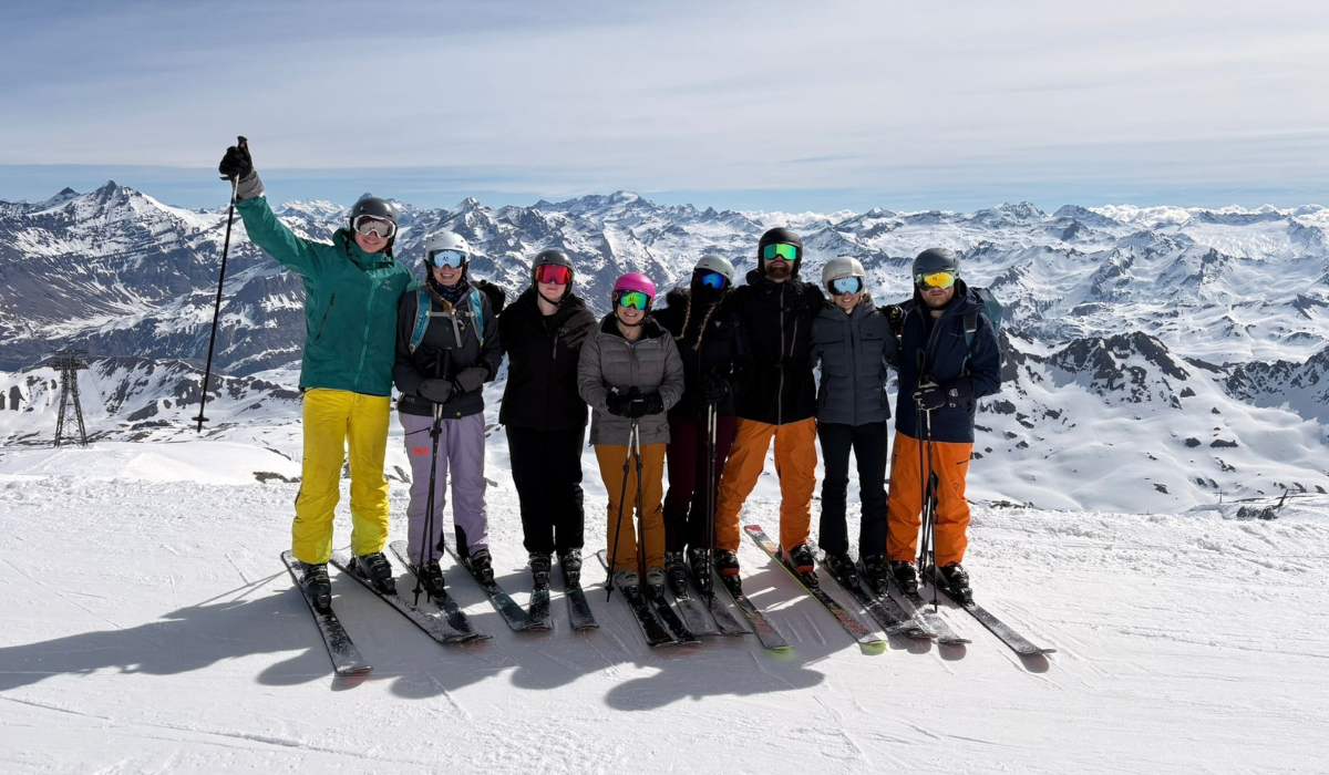 Line of skiiers standing shoulder to shoulder with snowy mountain peaks in the background.